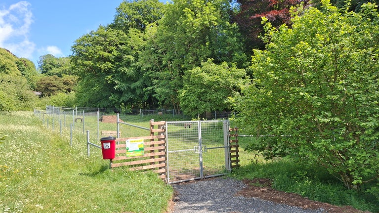A fenced off-lead area for dogs with agility activities inside and a sign and dog waste bin to the left of the metal entrance gates
